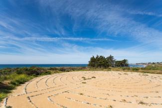 An aerial view of a labyrinth with intricate patterns, surrounded by greenery, the ocean on the horizon, in a serene outdoor setting. The photo captures the symmetry and design of the labyrinth, reflecting a peaceful and contemplative atmosphere.